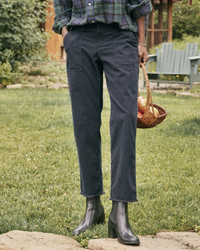 A person standing in a garden wearing Frank & Eileen's Italian Cotton Twill Blackrock Utility Pants in Washed Black, a plaid shirt, and black ankle boots, holding a basket with fruits.