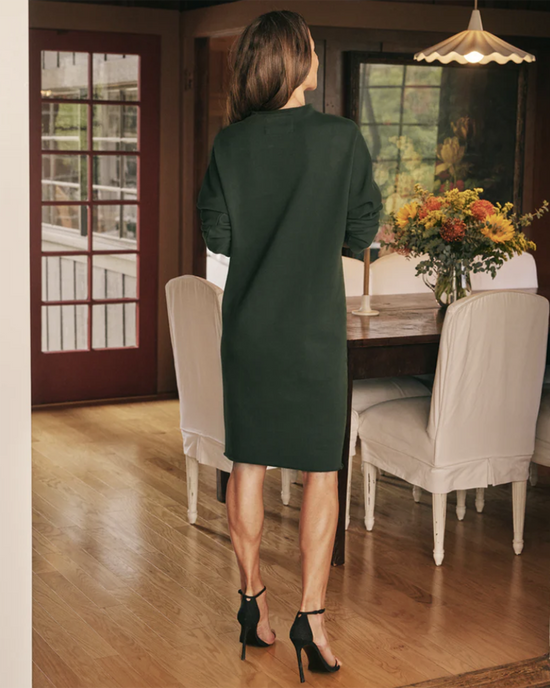 A woman in a dark green Frank & Eileen Izzie Funnel Neck Short Dress in Evergreen and black heels standing in a dining room facing away from the camera.
