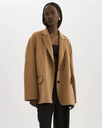 A woman wearing an oversized, single-breasted Lamarque Ennis Jacket in Camel over a black outfit against a white background.
