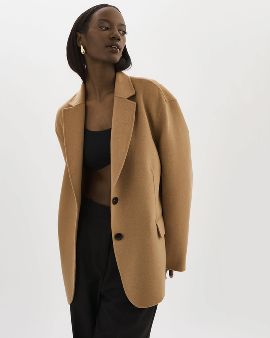 A woman in an oversized, single-breasted Ennis Jacket in Camel by Lamarque and black attire posing against a white background.