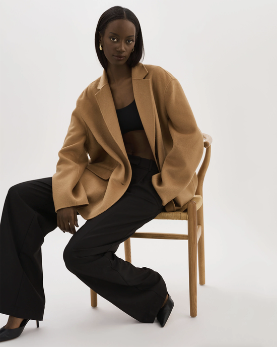 A woman sits on a chair modeling a Lamarque Ennis Jacket in Camel and black trousers.