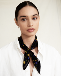 Woman wearing a Chan Luu Spring Floral Bandana in Black tied around her neck, paired with a white shirt and subtle makeup.