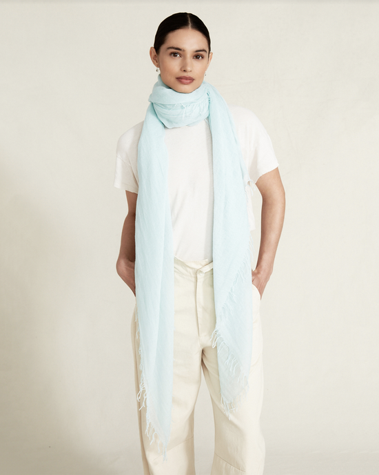 A woman in a white t-shirt and beige pants, wearing a Chan Luu Cashmere & Silk Scarf in Aqua with fringed trims, standing against a neutral background.