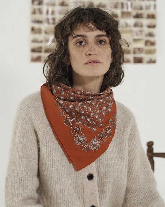 A person with curly hair wearing a beige sweater and a Mois Mont Bandana No 675 in Tomette around their neck, standing in front of a white wall with framed artworks.