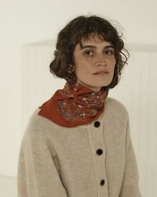 A person with curly hair wearing a beige cardigan and a Mois Mont Bandana No 675 in Tomette around their neck.