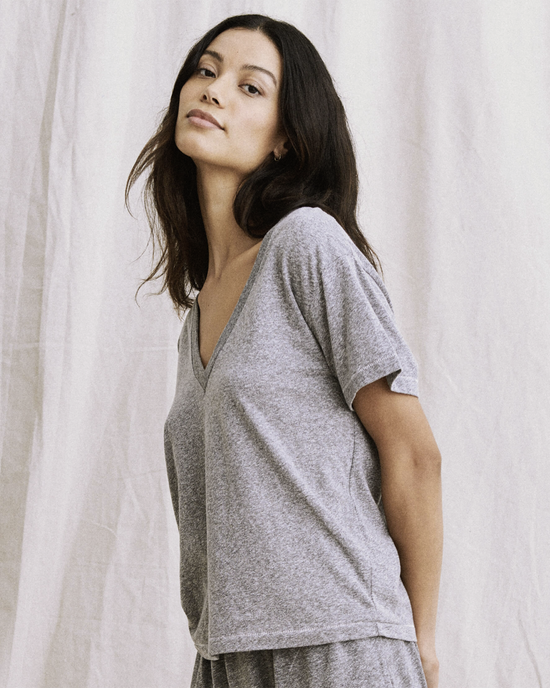 A woman in The V Neck in Heather Grey t-shirt by the Great with pitched sleeves stands against a light fabric background, looking at the camera with a subtle expression.