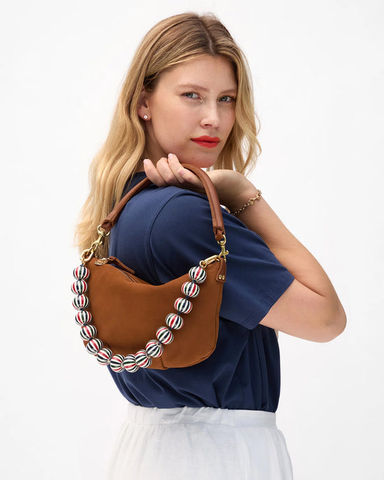 A woman with blonde hair, in a navy blue top and white skirt, carries the Clare V. Petit Moyen Messenger crossbody bag in Smooth Nubuck Hazelnut with a beaded strap. She looks at the camera against a plain background.