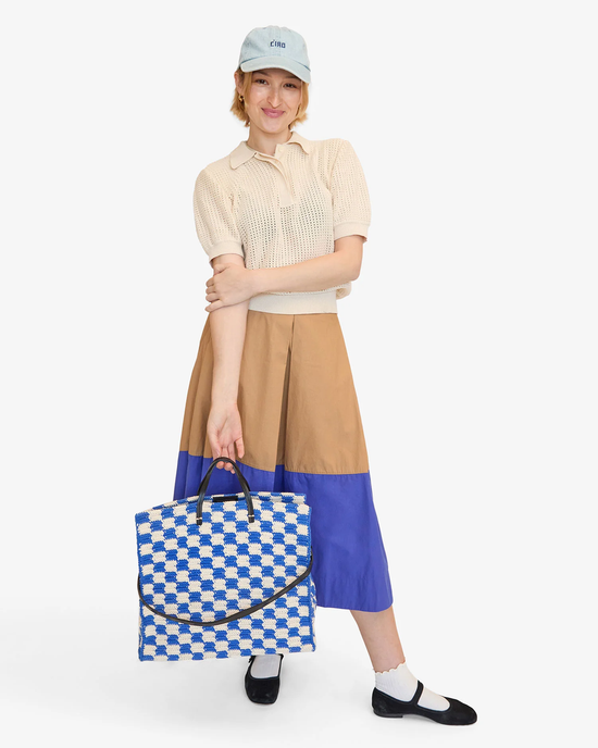 Woman in a color-blocked skirt and a beige polo shirt, wearing a cap and holding a Clare V. Summer Simple Tote in Cobalt & Cream Crochet, posing on a white background.