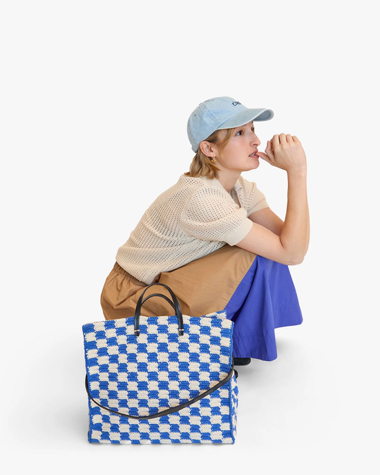 A person in a cap and casual outfit sitting and looking to the side with a Clare V. Summer Simple Tote in Cobalt & Cream Crochet next to them on a white background.