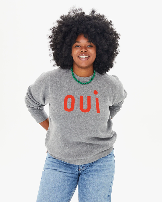 Woman smiling in a Clare V Oui Sweatshirt in Dark Heather Grey w/ Bright Poppy, paired with blue jeans and a green necklace.