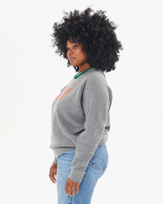 A woman with curly hair wearing a Clare V Oui Sweatshirt in Dark Heather Grey w/ Bright Poppy made in the USA and blue jeans, standing sideways and looking over her shoulder.
