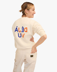 Woman in Clare V Flocked Deja Vu Sweatshirt in Cream w/ Cobalt & Zucca, with "deal uv" text on the back, smiling over her shoulder, wearing coordinating cargo pants.