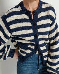 A person wears the Nation LTD Tessa Cardigan in Marine Stripe, a navy and white knit cotton cardigan with black buttons and an oversized fit, paired with blue jeans. They stand with one hand on their hip against a light background.