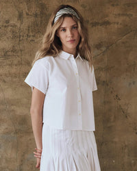 A woman with long hair and a striped headband, dressed in the Greats versatile staple, The Valor Top in White, confidently stands against a textured wall.