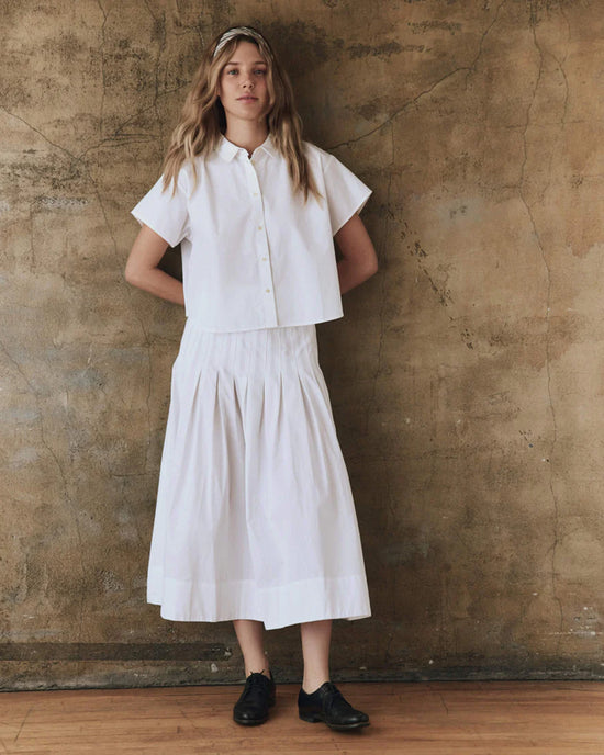 A person with long hair wears the versatile Valor Top in White by the Great, paired with a matching ankle-length skirt, standing against a textured brown wall and wooden floor.