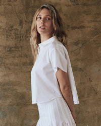 With long hair flowing, a person models The Valor Top in White by the Great against a textured brown backdro and casts a subtle glance backward.