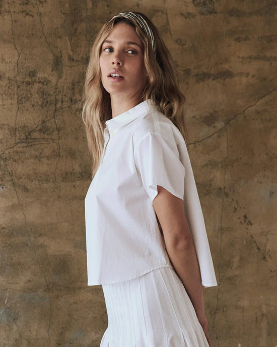 With long hair flowing, a person models The Valor Top in White by the Great against a textured brown backdro and casts a subtle glance backward.
