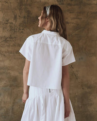 A woman with long hair wears The Valor Top in White by the Great and a white skirt, standing in front of a textured brown wall.
