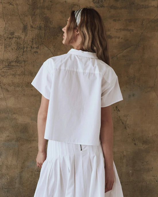A woman with long hair wears The Valor Top in White by the Great and a white skirt, standing in front of a textured brown wall.