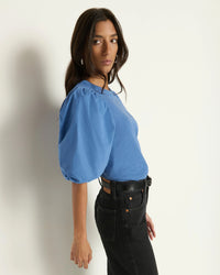 A woman with long brown hair stands sideways against a white wall, wearing the Nation LTD Tippi Top in Harbor Blue, tucked into high-waisted black jeans and accessorized with a black belt. She glances over her shoulder with a neutral expression.