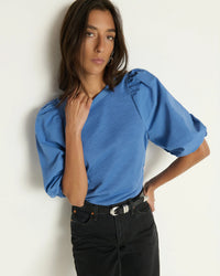 A woman with straight brown hair wears the Nation LTD Tippi Top in Harbor Blue and black jeans with a belt, posing against a white wall with one hand on her hip and the other touching her shoulder.