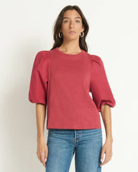 A woman wears the Nation LTD Tippi Top in Russet with blue jeans, standing against a plain light background and looking straight ahead with her arms at her sides.