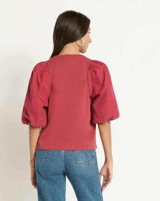 A woman with long brown hair is seen from behind, wearing the Nation LTD Tippi Top in Russet with puffed elbow-length sleeves and blue jeans. She accessorizes with a silver hoop earring and has one arm slightly bent.
