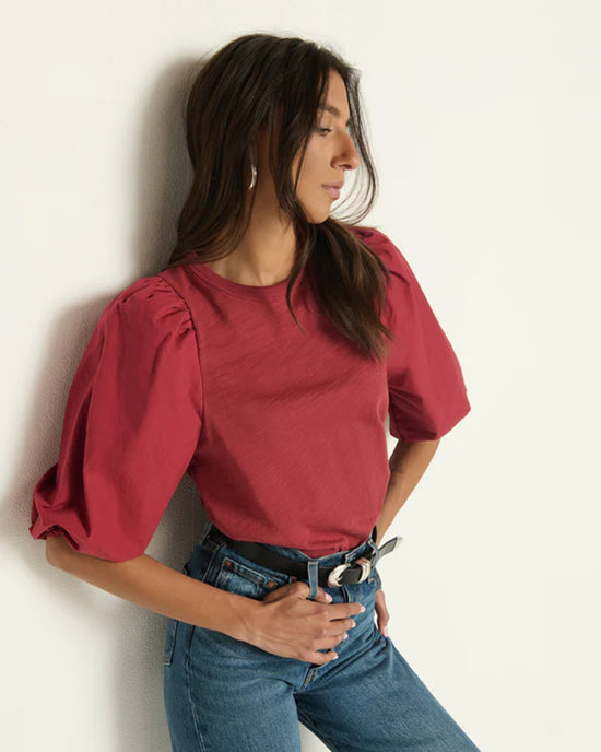 Woman with long brown hair wears the Nation LTD Tippi Top in Russet and blue jeans, standing with one hand in her pocket and leaning against a light-colored wall, gazing to the side.