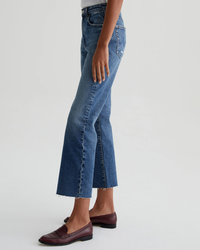 Lower half of a person wearing blue AG Jeans Kinsley in 15Ys Upstate in a cropped length and brown loafers standing against a neutral background.