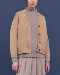 Woman wearing a Light Camel Inara Cardigan by Demylee over a turtleneck with a pleated skirt against a blue background.