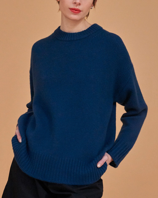 Woman in a Demylee Rell Sweater in Teal Blue and black pants against a tan background.