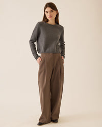 A woman with straight brown hair stands against a beige background wearing the Demylee Melina Cotton Cashmere Sweater in Dark Heather Grey, paired with high-waisted, wide-leg brown trousers and black shoes. Her left hand is in her pocket.