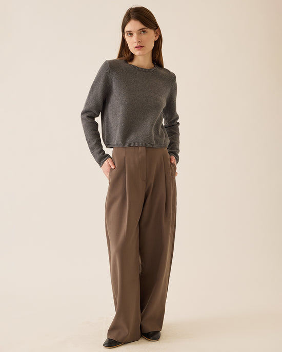 A woman with straight brown hair stands against a beige background wearing the Demylee Melina Cotton Cashmere Sweater in Dark Heather Grey, paired with high-waisted, wide-leg brown trousers and black shoes. Her left hand is in her pocket.