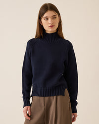 A young woman with straight brown hair wears the Demylee Kaitlyn Cotton Cashmere Sweater in Navy and loose brown trousers, standing against a plain beige background and looking slightly to her left.