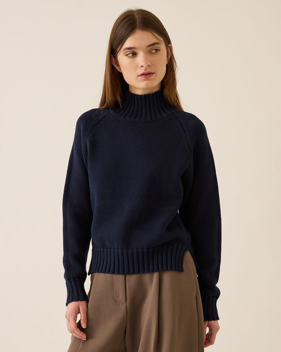 A young woman with straight brown hair wears the Demylee Kaitlyn Cotton Cashmere Sweater in Navy and loose brown trousers, standing against a plain beige background and looking slightly to her left.