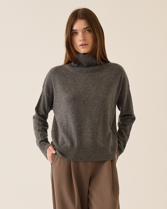 A woman with straight brown hair wears the Demylee Gianna Cashmere Turtleneck in Dark Heather Grey and brown trousers, standing against a plain beige background with her left hand in her pocket.