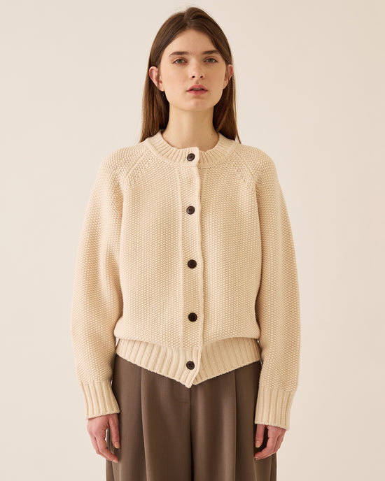 A woman with straight brown hair wears the Demylee Davi Merino Wool Cardigan in Off White, featuring dark buttons and wide ribbed cuffs, paired with loose brown pants, standing against a plain light background.
