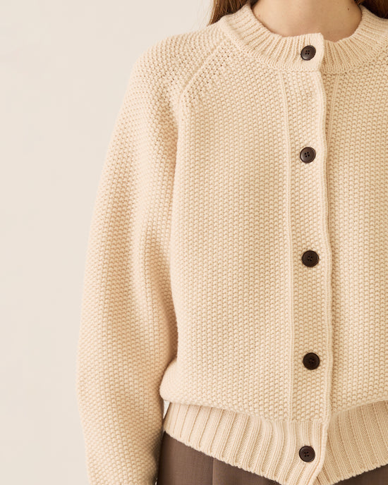 A person wears the Demylee Davi Merino Wool Cardigan in Off White with dark buttons, paired with brown pants. The cropped image shows only their lower face and torso, from shoulders to mid-thigh.