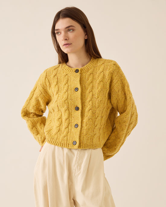 A woman with long brown hair wears the Demylee Kinsley Speckled Wool Cardigan in Corn, buttoned and paired with high-waisted cream trousers, standing against a plain beige background.