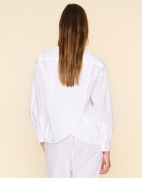 Rear view of a person with long hair wearing a white XiRENA Emile Top with a Collared V Neck and light-colored pants against a beige background.
