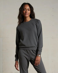 A woman with long wavy hair smiles softly at the camera, wearing the perfectwhitetee Alexi French Terry Sweatshirt in Charcoal and matching sweatpants, standing against a plain light-colored wall.