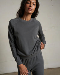 A person in a perfectwhitetee Alexi French Terry Sweatshirt in Charcoal with matching sweatpants stands indoors against a beige wall, confidently looking at the camera and pulling slightly at the sweatshirt's hem.