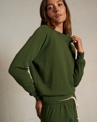 A person wearing the Alexi French Terry Sweatshirt in Fern Green by perfectwhitetee and matching sweatpants stands against a plain, light background, holding the sweatshirt’s collar and looking at the camera.