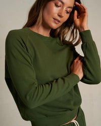 A woman in a Fern Green Alexi French Terry Sweatshirt by perfectwhitetee with matching pants stands against a neutral background, touching her hair and looking down gently.