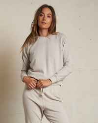 A woman with long brown hair wears the perfectwhitetee Alexi French Terry Sweatshirt in Silver, standing confidently against a plain light wall and looking at the camera.