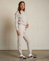 A woman stands indoors in the perfectwhitetee Alexi French Terry Sweatshirt in Silver with matching joggers, white socks, and black Nike sneakers, posing by a plain light-colored wall with one hand at her side.