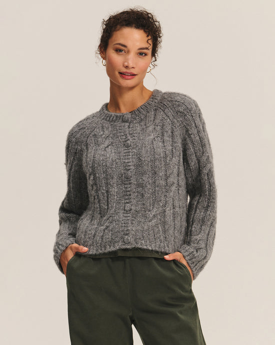 A woman with curly hair poses with her hands in her pockets, wearing the Velvet by Graham & Spencer Allison Long Sleeve Crew Cardi in Heather Grey and dark green pants against a plain light background.