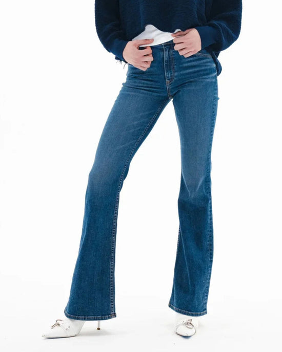 Wearing the ASKK NY Bond Street in Paris mid-rise, dark blue flare jeans made from Japanese stretch denim, a person stands against a white background with their hands near the waistband, paired with a navy sweater and white heeled shoes.