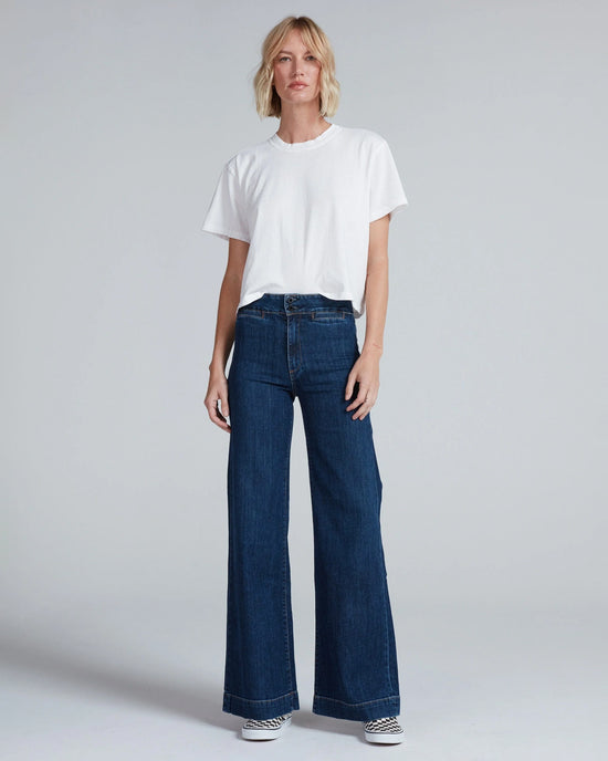 A person stands facing forward against a light gray background, wearing a plain white t-shirt, ASKK NY Brighton in Dark Stone high rise wide leg jeans made from Japanese stretch denim, and striped slip-on shoes.