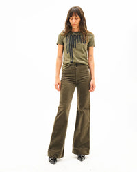 A woman stands against a white background wearing an olive green graphic tee, black pointed-toe shoes, and ASKK NY Brighton Corduroy Wide Leg pants in Safari Green. She looks down with her hands at her sides.